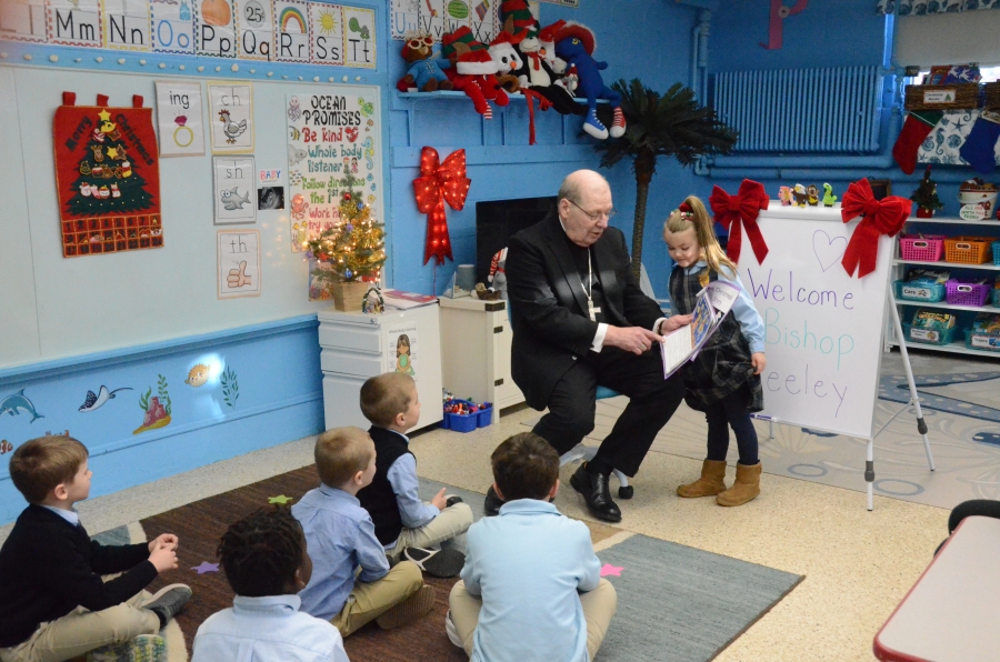 A student reads a page of a book to the bishop as other students seated look on 