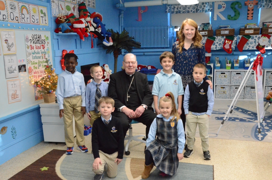 A group of six students and a teacher surround a seated bishop 