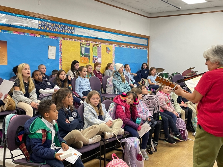 Group of children in chair singing, woman at front of the room with a guitar