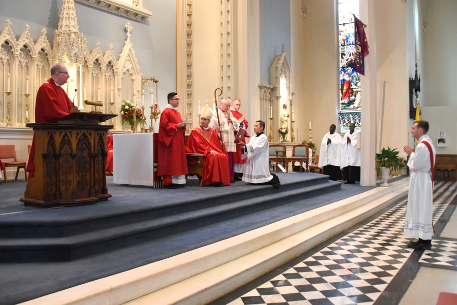 Deacon Matthew Valles stands at the foot of the stairs, while Msgr. Andrew Dubois testifies to his readiness.